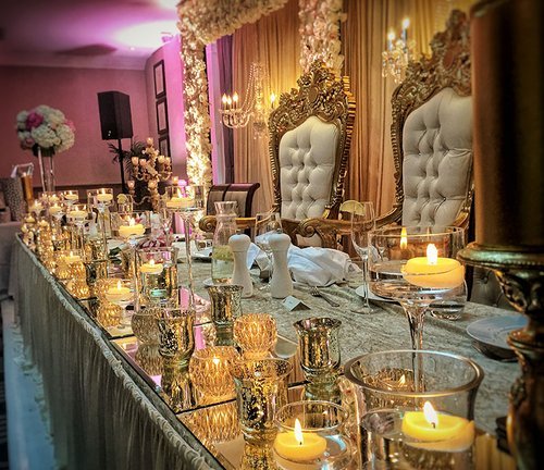 A mirrored top table lined with gold candle holders and crystal glassware, with carved ivory throne chairs