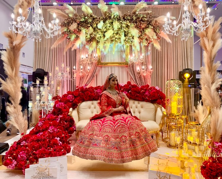 A bridal stage edged with red roses and pampas grass beneath a suspended floral installation and crystal chandeliers